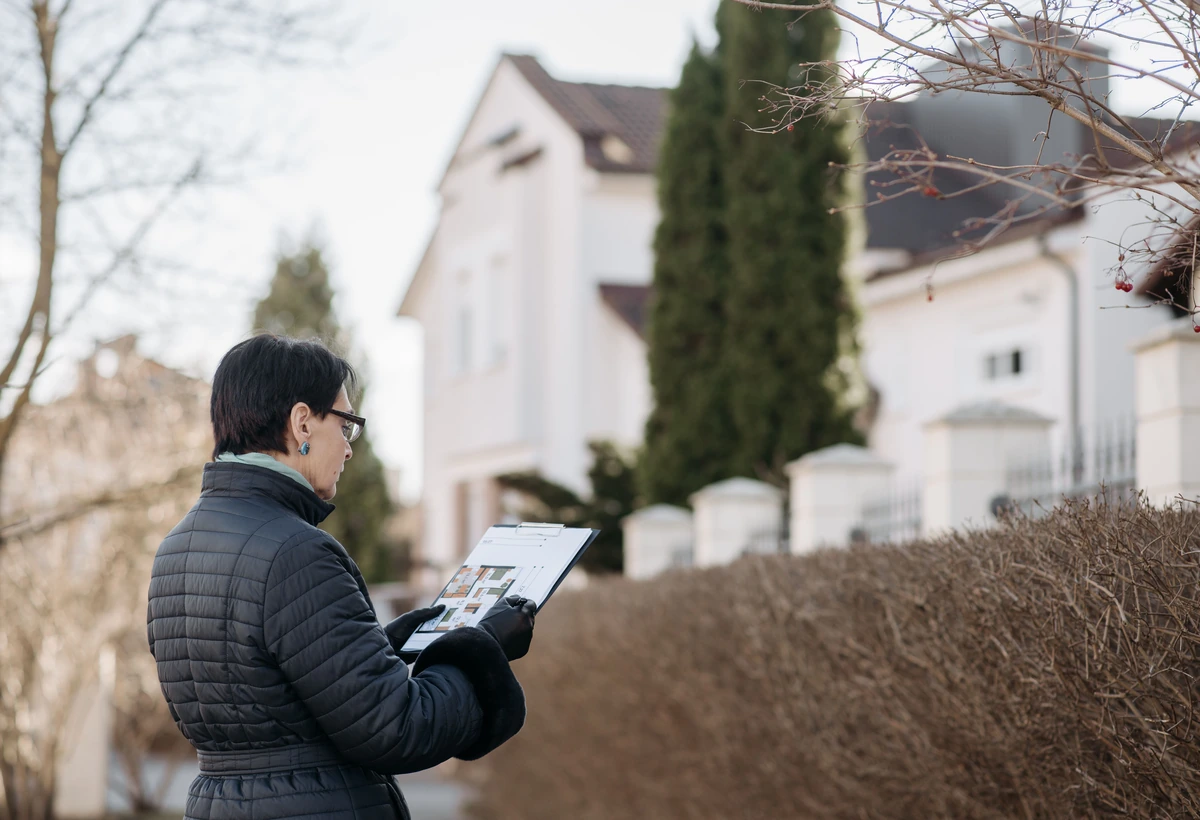 Immobilienfachkraft betrachtet ein Wohnhaus und hält Objektunterlagen bereit